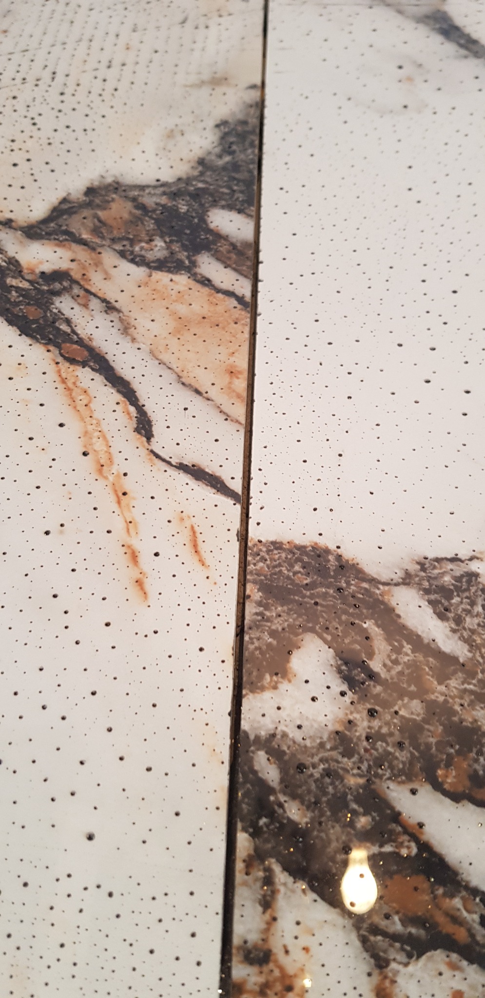 Dwie płytki podłogowe z białego marmuru z brązowymi i czarnymi żyłami, pokryte drobnymi, ciemnymi plamkami, ułożone obok siebie z widoczną fugą.
