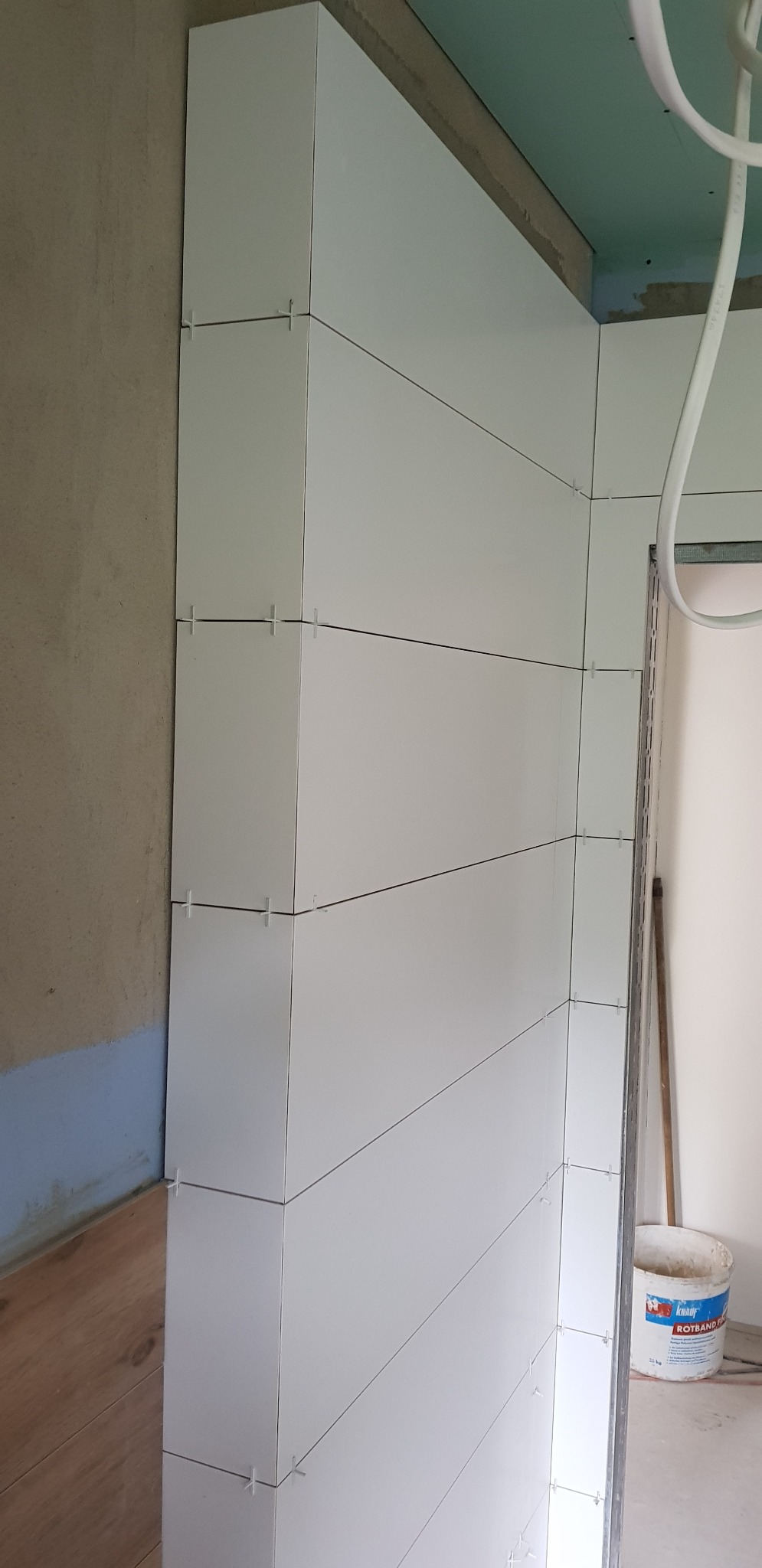 Ściana w trakcie układania białych, prostokątnych płytek z widocznymi krzyżykami dystansowymi, fragment surowej ściany i podłogi, oraz wiadro z zaprawą.