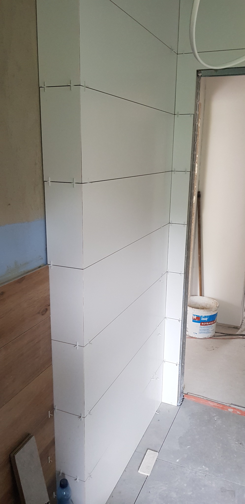 Fragment ściany z białych, prostokątnych płytek, ułożonych poziomo z użyciem krzyżyków dystansowych, widoczne niedokończone prace i wiadro z zaprawą w tle.