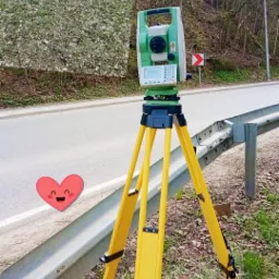 Pomiar geodezyjny w terenie: teodolit na żółtym statywie ustawiony przy drodze, w tle pracująca osoba i dom na wzgórzu.
