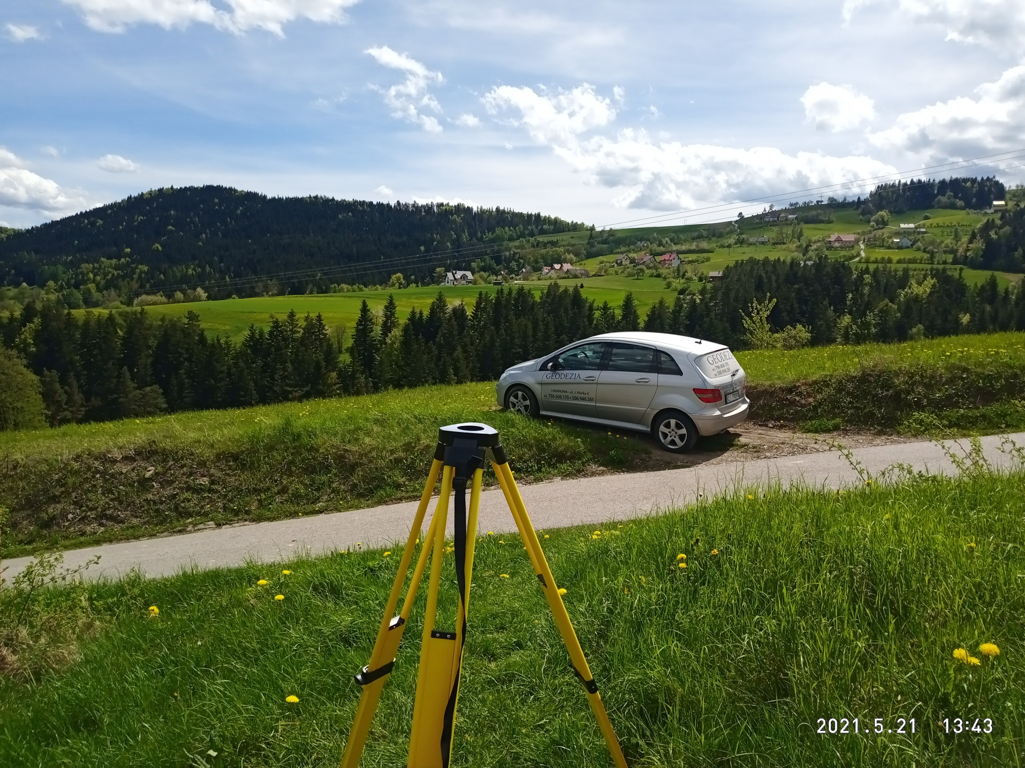 Żółty statyw geodezyjny na tle malowniczego krajobrazu z zielonymi wzgórzami, lasami i srebrnym samochodem z napisem 'GEODEZJA' w tle, błękitne niebo z chmurami.