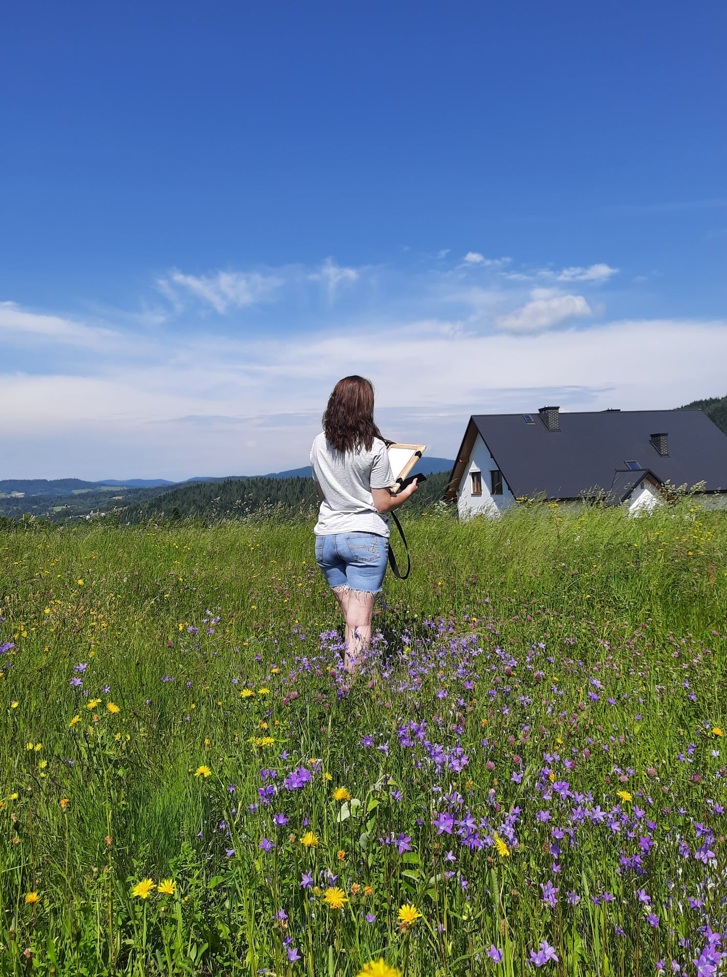 Kobieta z brązowymi włosami, ubrana w dżinsowe spodenki i białą koszulkę, stoi tyłem na łące pełnej fioletowych i żółtych kwiatów, trzymając w rękach deskę kreślarską; w tle dom z ciemnym dachem...