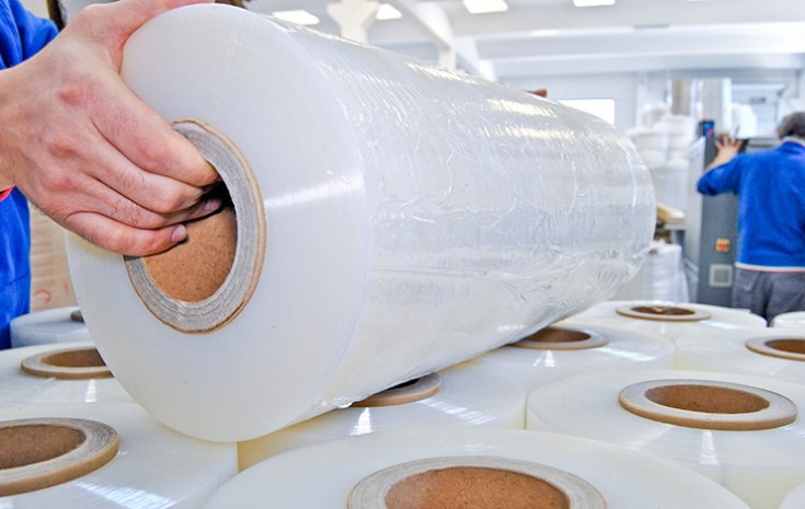 Ręka pracownika trzymająca dużą rolkę przezroczystej folii stretch na tle stosu identycznych rolek w hali produkcyjnej.