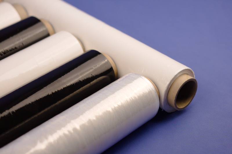 Rzędy rolek folii stretch w kolorach białym i czarnym, ułożone na niebieskim tle z delikatną teksturą.