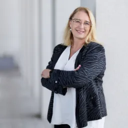 Uśmiechnięta blondynka w okularach, oparta o białą kolumnę, ubrana w białą bluzkę i czarną kurtkę w zebrę, skrzyżowane ręce, profesjonalny portret.