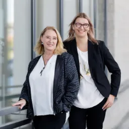 Portret dwóch uśmiechniętych kobiet w biznesowych strojach, stojących na tle nowoczesnego budynku z przeszkloną fasadą.