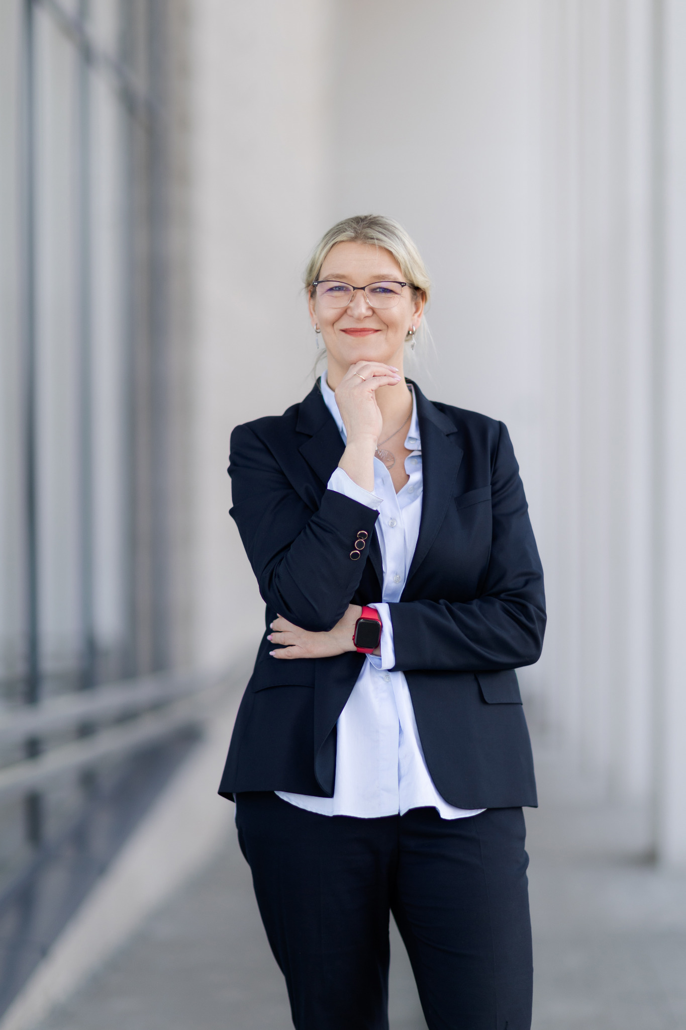 Portret kobiety w okularach i granatowym garniturze, z ręką pod brodą, na tle jasnych kolumn budynku.