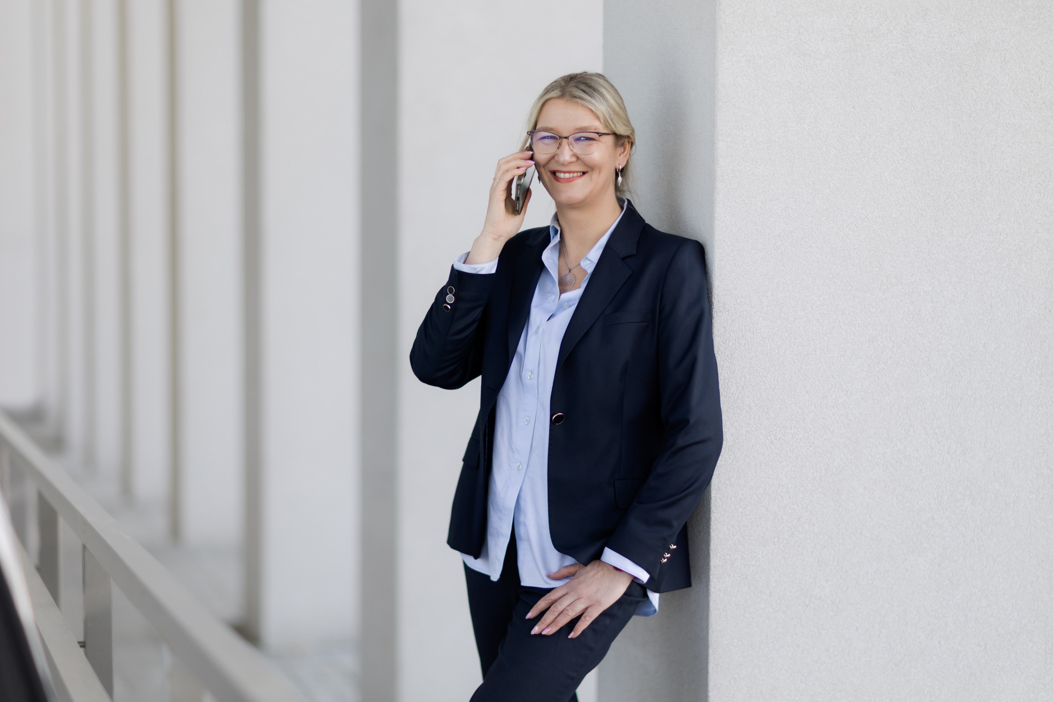 Uśmiechnięta blondynka w okularach, ubrana w granatowy żakiet i jasną koszulę, rozmawia przez telefon oparta o białą kolumnę.