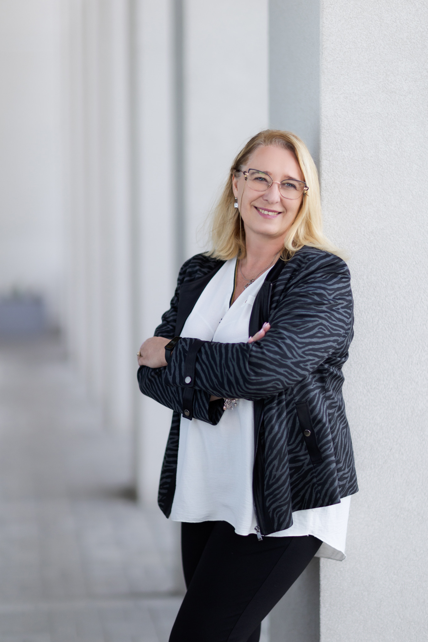 Uśmiechnięta blondynka w okularach, oparta o białą kolumnę, ubrana w białą bluzkę i czarną kurtkę w zebrę, skrzyżowane ręce, profesjonalny portret.