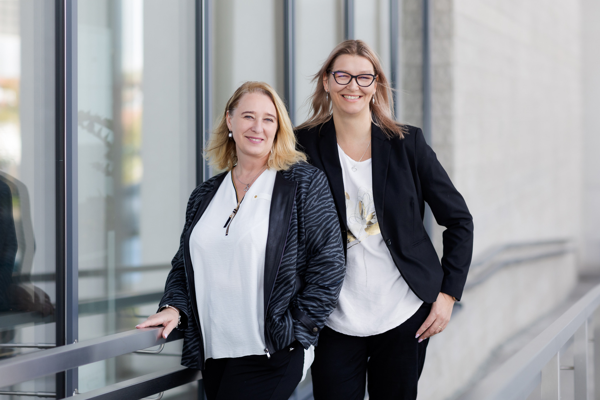 Portret dwóch uśmiechniętych kobiet w biznesowych strojach, stojących na tle nowoczesnego budynku z przeszkloną fasadą.