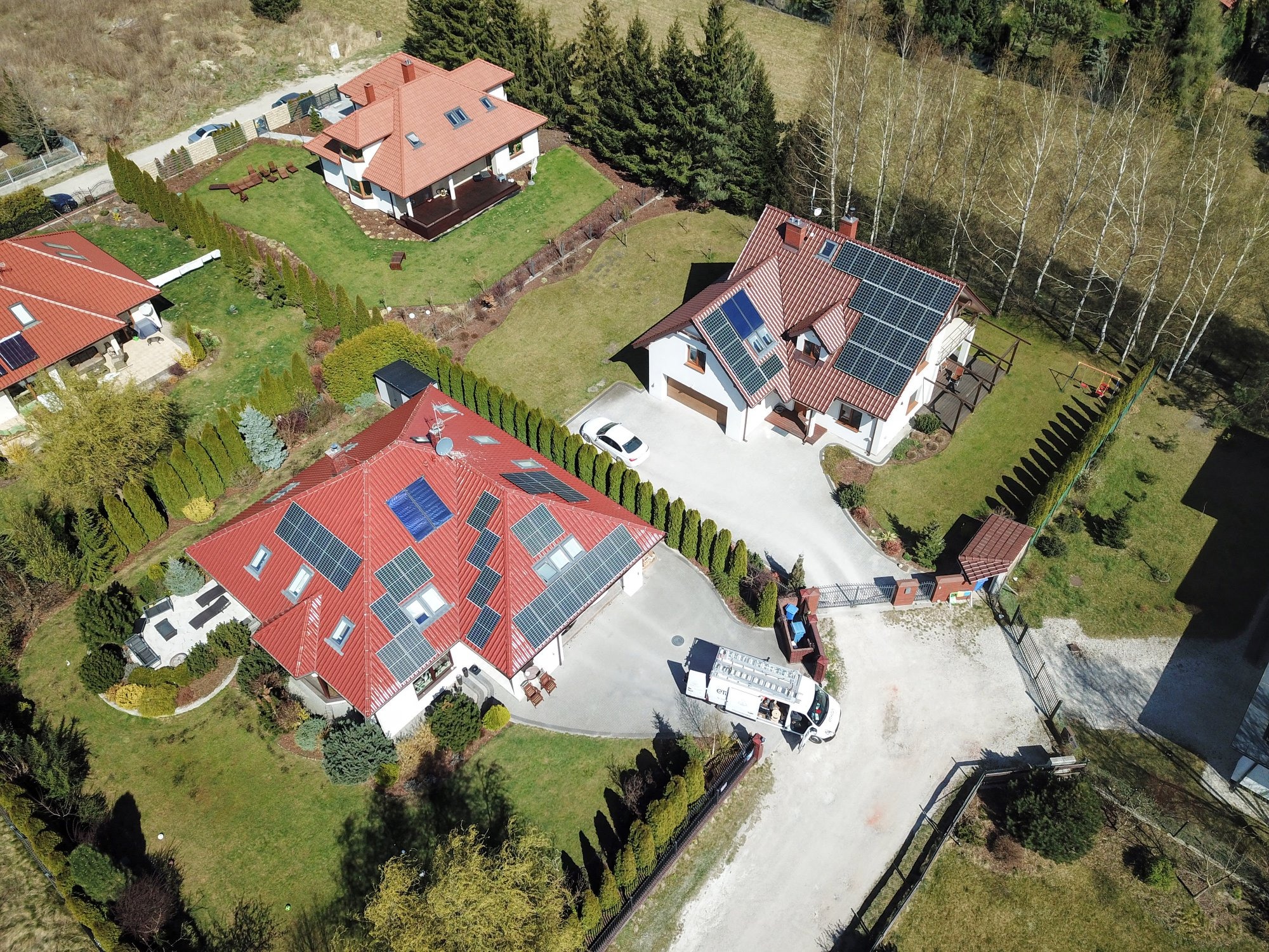 Widok z lotu ptaka na trzy domy jednorodzinne z panelami słonecznymi na dachach; jeden z domów ma zaparkowany samochód serwisowy z drabiną.