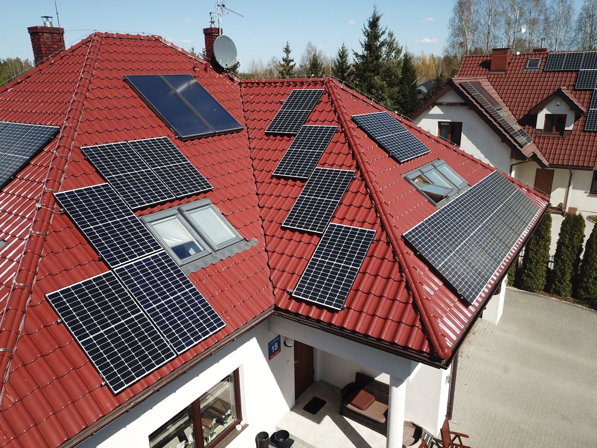 Czerwony dach z dachówki ceramicznej pokryty panelami słonecznymi; widoczne okna dachowe, antena satelitarna i numer domu na ścianie.