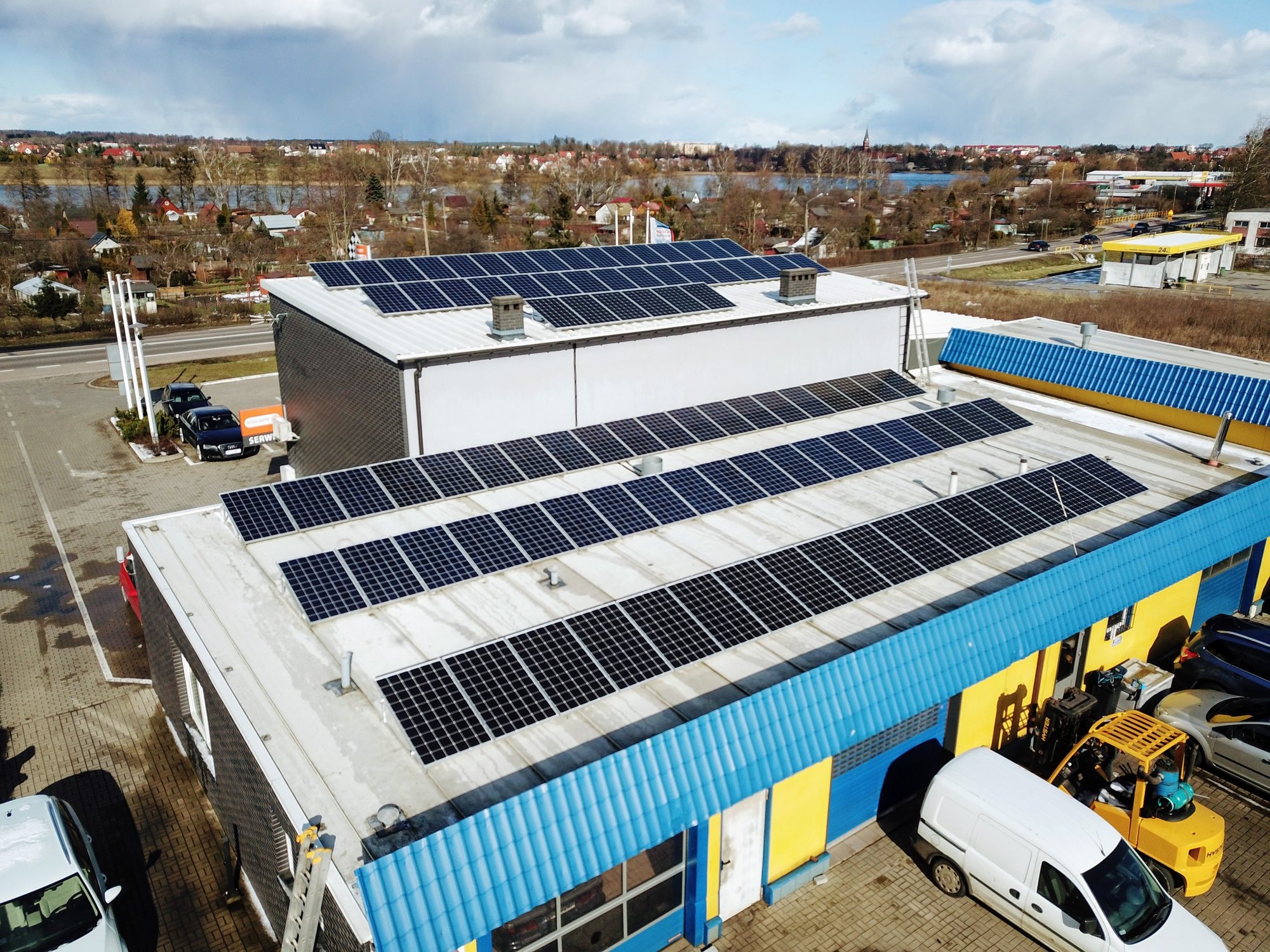 Widok z góry na budynek z panelami słonecznymi na dachu, w tle jezioro i zabudowania mieszkalne, na dole zaparkowane samochody i wózek widłowy.