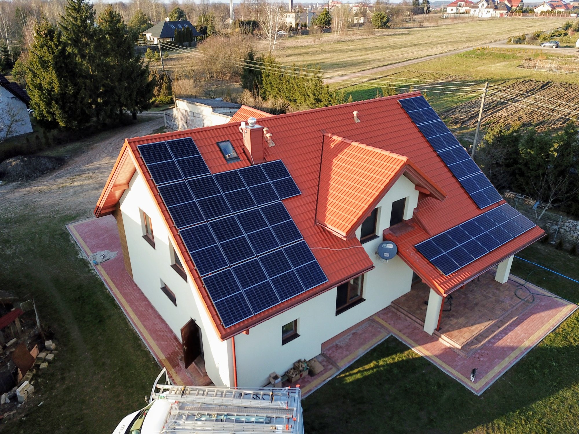 Widok z drona na dom jednorodzinny z czerwoną dachówką i panelami słonecznymi na dachu, drabina oparta o ścianę, otoczony zielenią i polami.