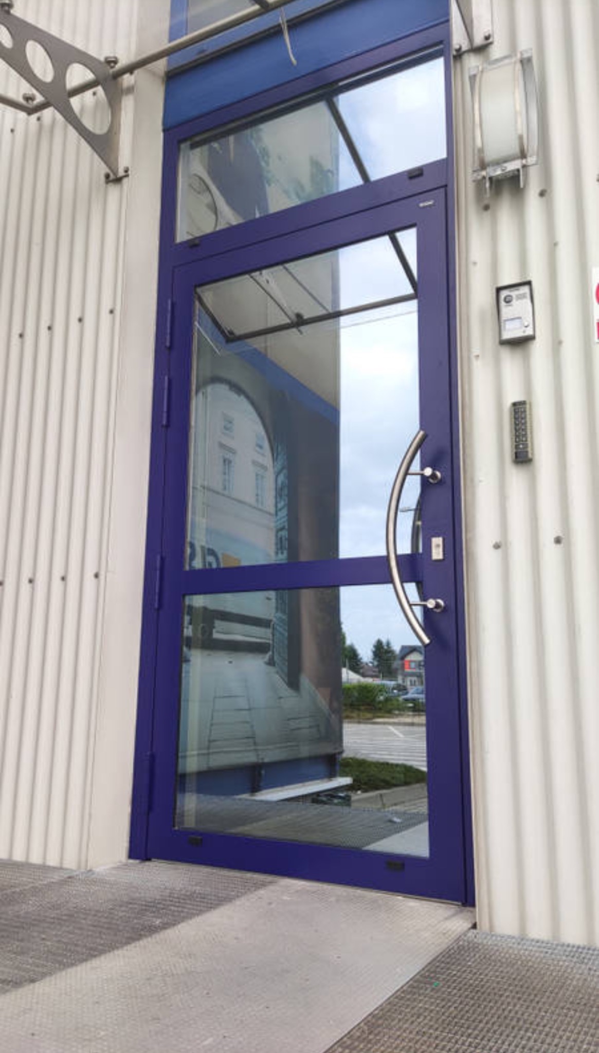 Niebieskie, przeszklone drzwi zewnętrzne z podłużnym uchwytem na tle białej, blaszanej elewacji z lampą i panelem numerycznym.