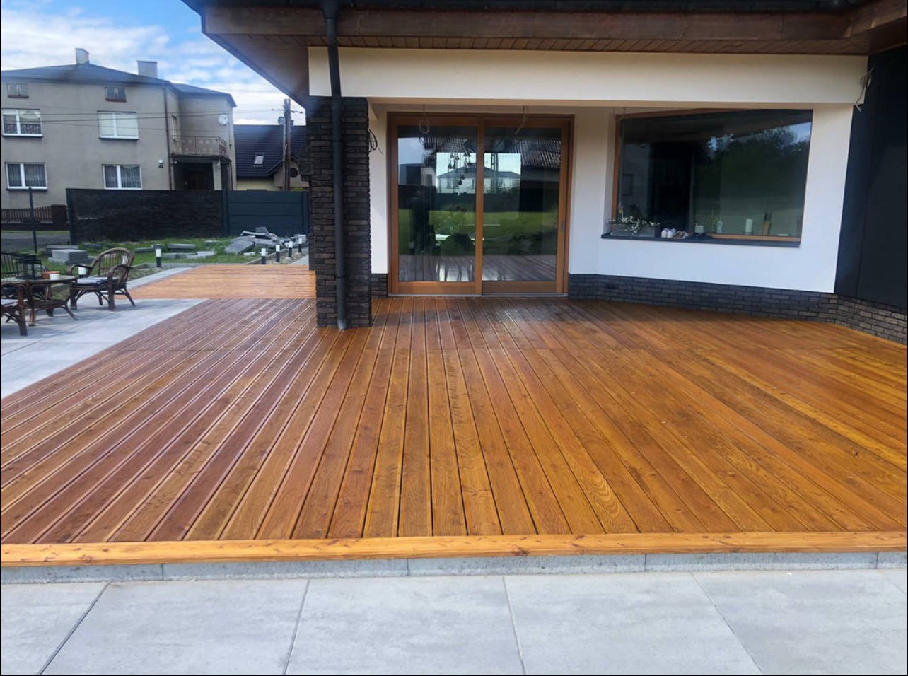 Drewniany taras z oknem HS i przesuwnymi drzwiami tarasowymi, wykończony cegłą klinkierową. Na tarasie widoczne meble ogrodowe.