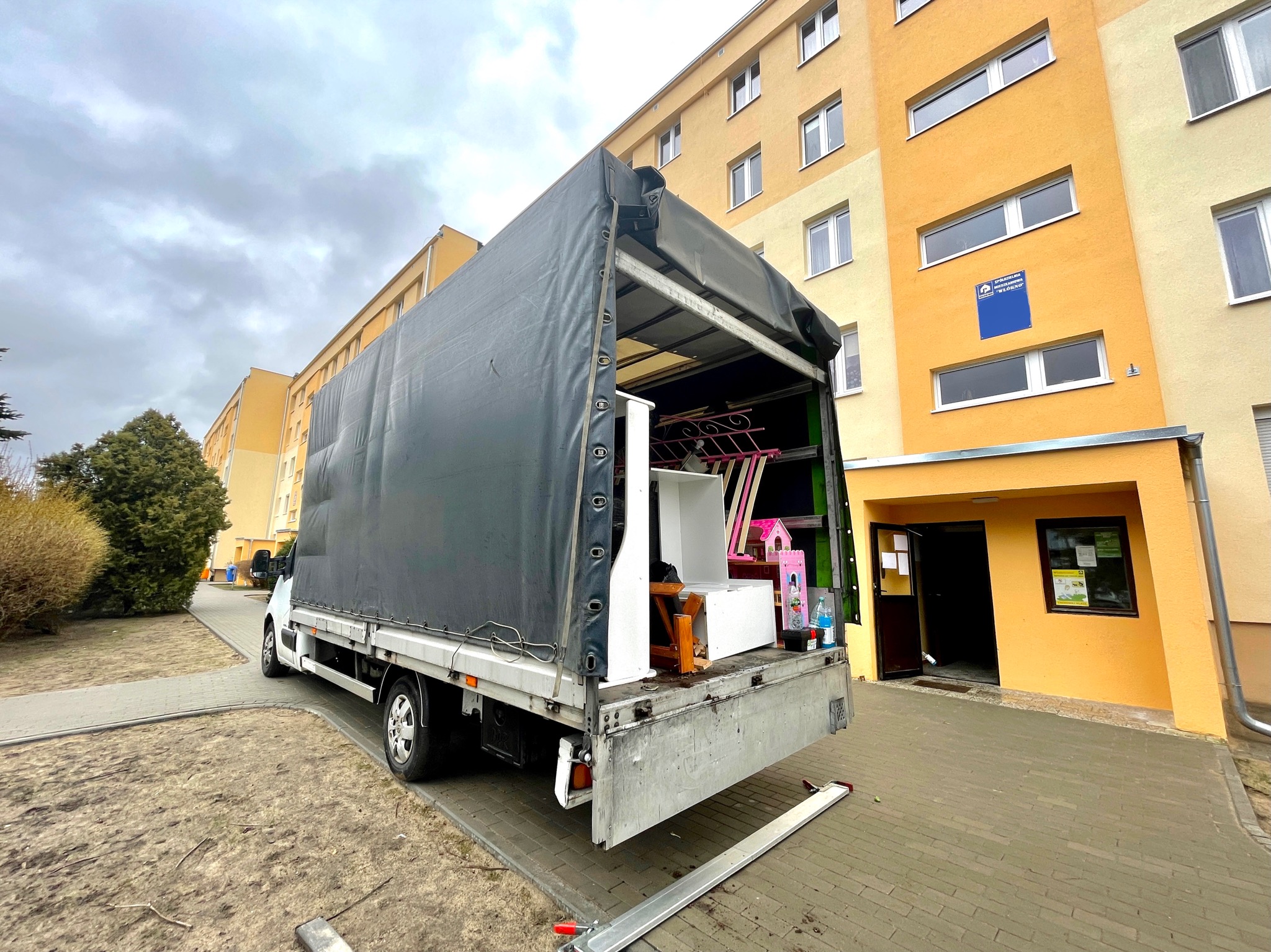 Otwarta paka samochodu dostawczego załadowana meblami i przedmiotami, rampa wysunięta na chodnik przed budynkiem mieszkalnym.