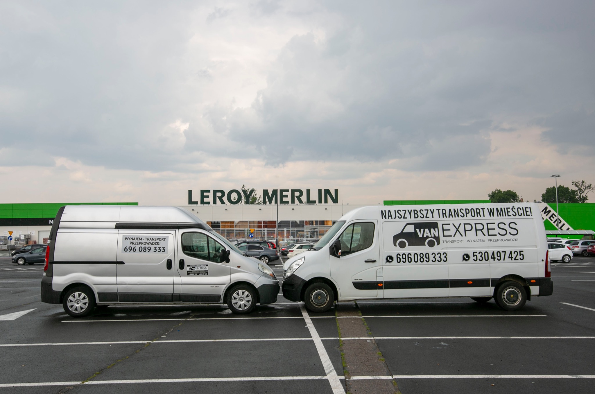 Dwa busy firm transportowych, srebrny i biały, zaparkowane na parkingu przed sklepem Leroy Merlin, z widocznymi numerami telefonów i hasłami reklamowymi na karoseriach.