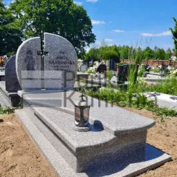 Jasnoszary, granitowy nagrobek z dwiema pionowymi płytami w kształcie półkola tworzącymi całość, z wizerunkiem postaci i wyrytym krzyżem; obok lampion na tle cmentarza.
