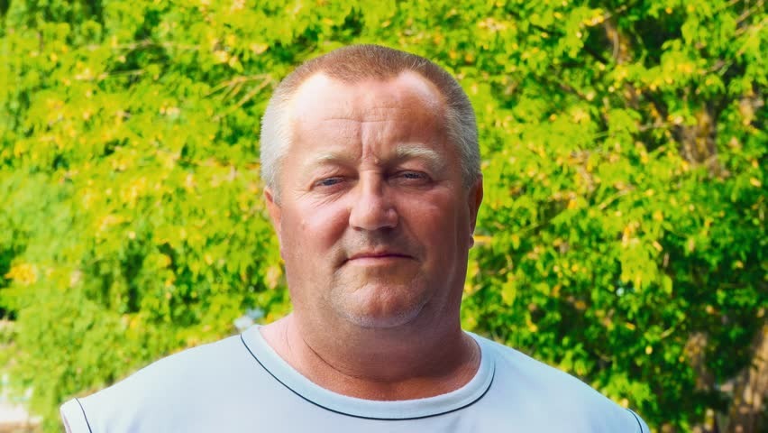 Portret mężczyzny na tle zielonego drzewa, prawdopodobnie właściciel firmy, w jasnej koszulce z ciemnym wykończeniem dekoltu, naturalne światło.