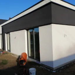 Nowoczesny dom jednorodzinny z białą elewacją i czarnym pasem na wysokości okien, widoczny nagrzewnica budowlana i resztki materiałów budowlanych na ziemi.