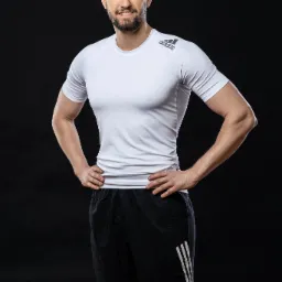 Uśmiechnięty mężczyzna w sportowym stroju marki Adidas, pozujący na czarnym tle, z rękami opartymi na biodrach.
