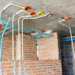 Białe rury instalacji elektrycznej prowadzone po suficie w surowym stanie, widoczne pomarańczowe puszki instalacyjne, fragment ściany z czerwonej cegły i niebieskie rury instalacji wodnej.