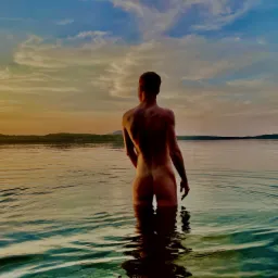 Mężczyzna stojący w wodzie po pas, widok z tyłu, w tle zachodzące słońce odbijające się w tafli jeziora.