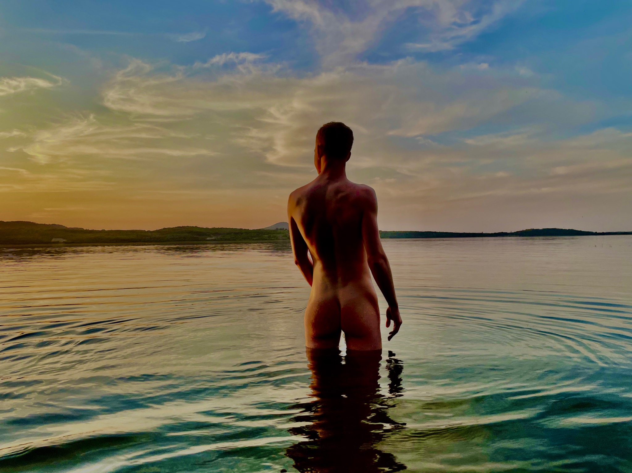 Mężczyzna stojący w wodzie po pas, widok z tyłu, w tle zachodzące słońce odbijające się w tafli jeziora.