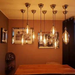 Grupa dziewięciu lamp wiszących z żarówkami Edisona, zwisających nad drewnianym stołem w jadalni z brązowymi ścianami i lustrzanymi obrazami.