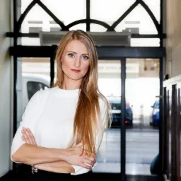 Kobieta z blond włosami w białej bluzce, skrzyżowane ręce, w tle przeszklone drzwi wejściowe do budynku.