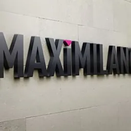 Czarny, przestrzenny napis 'MAXIMILANO' umieszczony na teksturowanej, jasnej ścianie. Nad literą 'I' widoczny mały, różowy trójkąt.