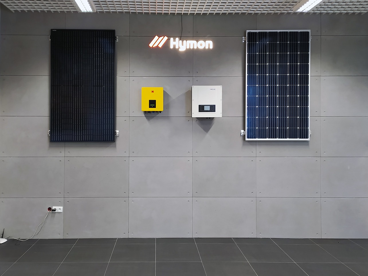 Prezentacja paneli fotowoltaicznych i falowników na szarej ścianie z płytek, oświetlonych górnym światłem, z widocznym logo Hymoń.