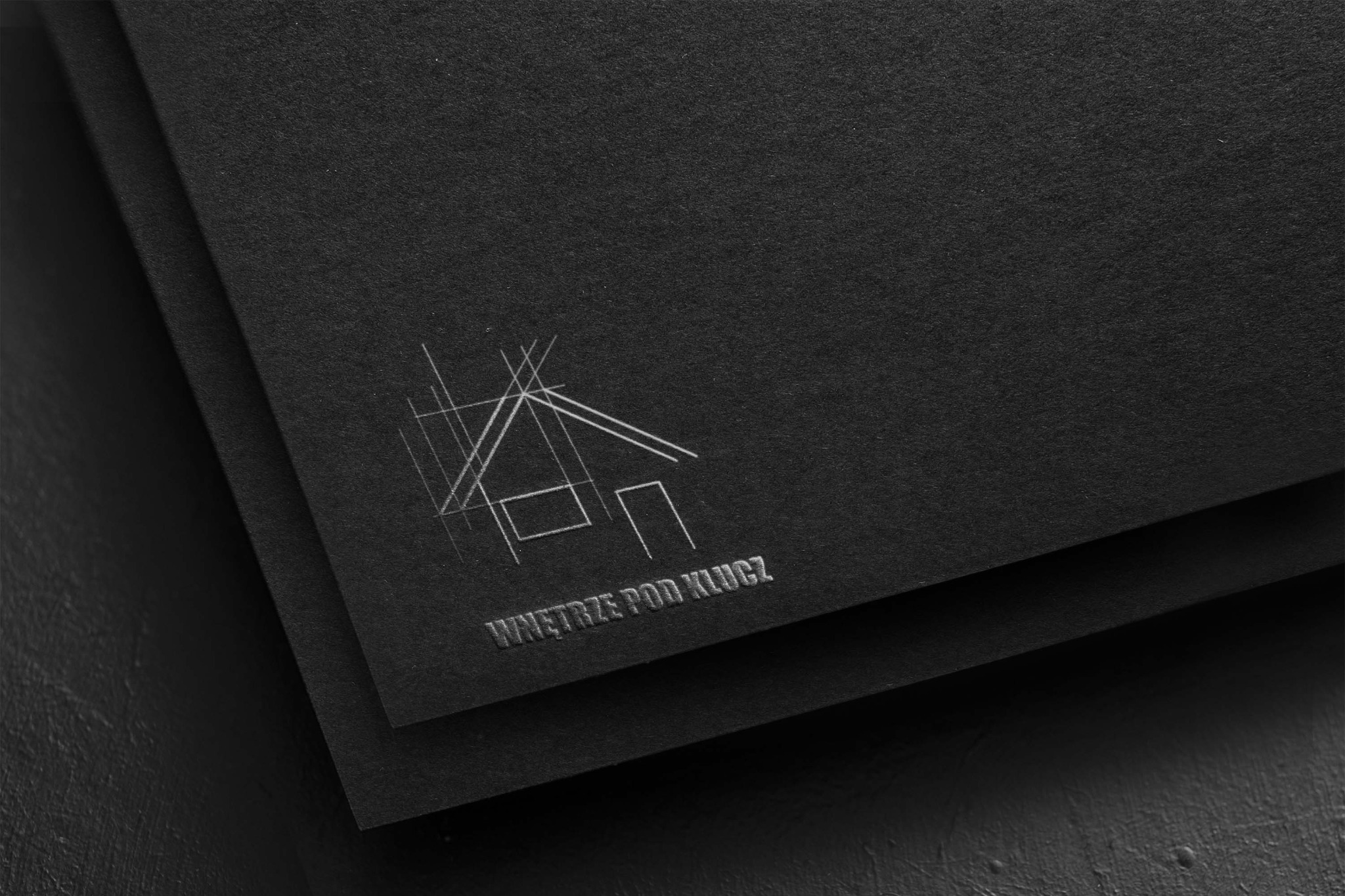 Czarny papier z wytłoczonym srebrnym logo firmy 'Wnętrze pod klucz' przedstawiającym schematyczny zarys domu.
