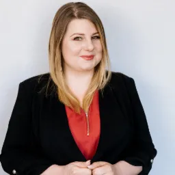 Portret uśmiechniętej kobiety w czarnej marynarce i czerwonej bluzce na jasnym tle.