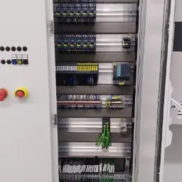 Otwarta szafa sterownicza z widocznymi elementami automatyki: przekaźniki, sterowniki PLC, okablowanie i przyciski awaryjne na panelu zewnętrznym.