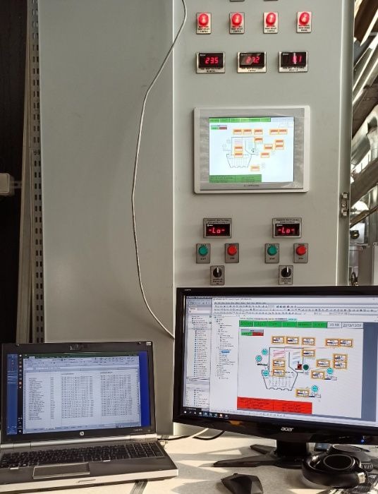 Panel sterowania z wyświetlaczami, przy którym stoją dwa laptopy z uruchomionym oprogramowaniem do monitoringu i sterowania procesem przemysłowym.