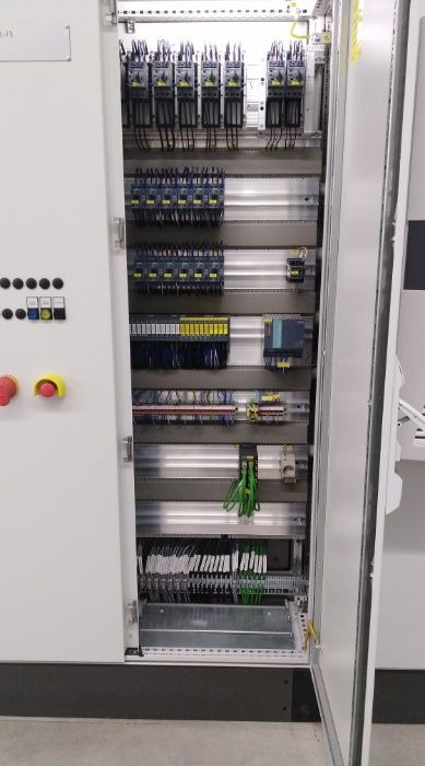 Otwarta szafa sterownicza z widocznymi elementami automatyki: przekaźniki, sterowniki PLC, okablowanie i przyciski awaryjne na panelu zewnętrznym.