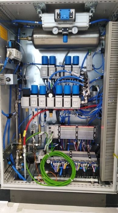 Wnętrze szafy sterowniczej z pneumatyką, widoczne liczne przewody, zawory, manometry i sterowniki PLC, z zielonym kablem zwiniętym w krąg na dole.