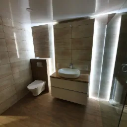 Nowoczesna łazienka z beżowymi płytkami, podświetleniem LED w ścianach i drewnopodobną podłogą, umywalka nablatowa na szafce z szufladami i toaleta w zabudowie.