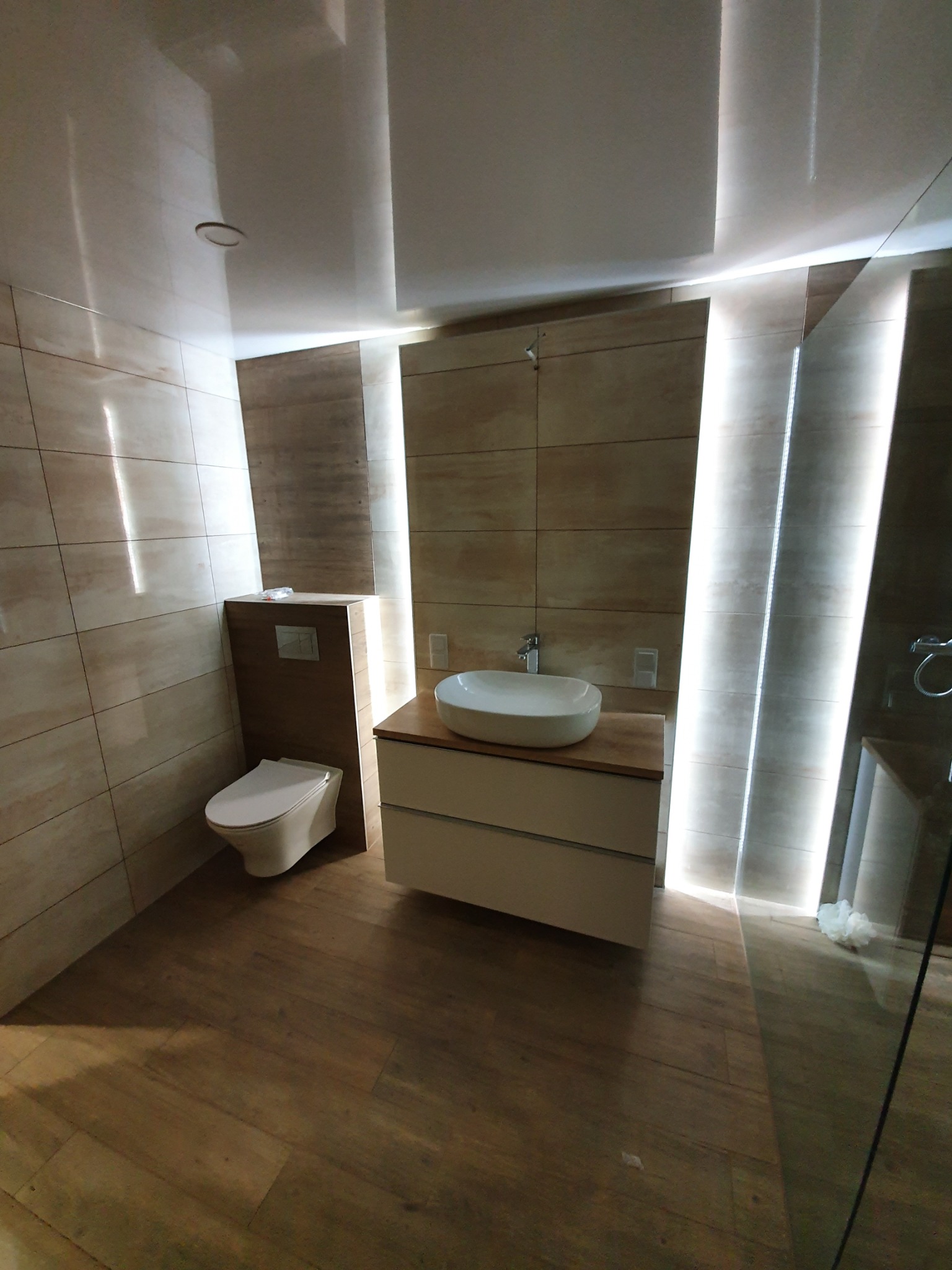 Nowoczesna łazienka z beżowymi płytkami, podświetleniem LED w ścianach i drewnopodobną podłogą, umywalka nablatowa na szafce z szufladami i toaleta w zabudowie.