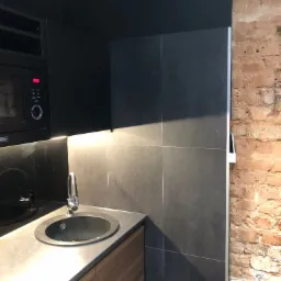 Czarny aneks kuchenny z okrągłym zlewem, baterią, mikrofalówką i fragmentem ceglanej ściany