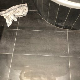 TiT Mariusz Bogusz - Czyszczenie ciemnych płytek podłogowych w łazience, widoczny ręcznik w kratę, ślady po wodzie oraz fragment wanny.