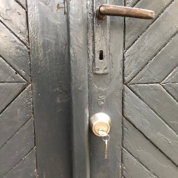 TiT Mariusz Bogusz - Szczegółowe ujęcie pomalowanych na szaro drzwi z wzorem w jodełkę, z zamontowanym nowym zamkiem cylindrycznym, w którym tkwią klucze, oraz starą, rdzewiejącą klamką.