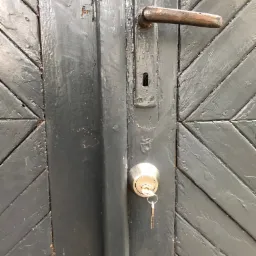 Szczegółowe ujęcie pomalowanych na szaro drzwi z wzorem w jodełkę, z zamontowanym nowym zamkiem cylindrycznym, w którym tkwią klucze, oraz starą, rdzewiejącą klamką.