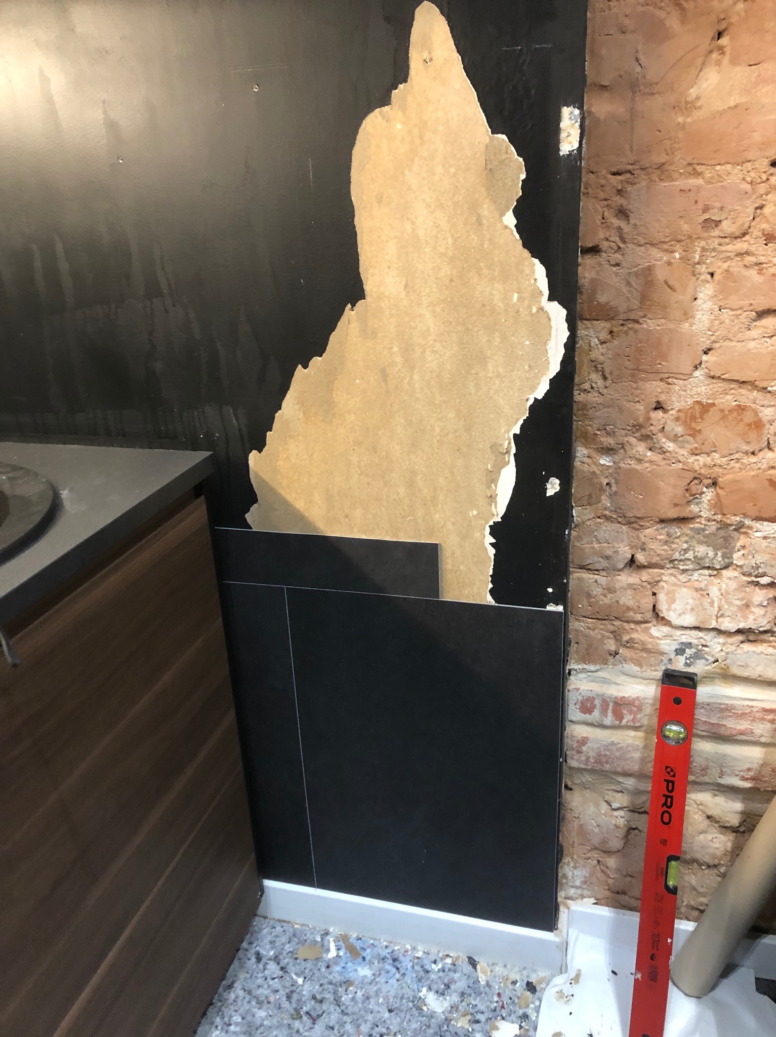 Układanie czarnych płytek ściennych w pomieszczeniu z odsłoniętą cegłą, widoczne resztki starej tapety i poziomica przy ścianie.