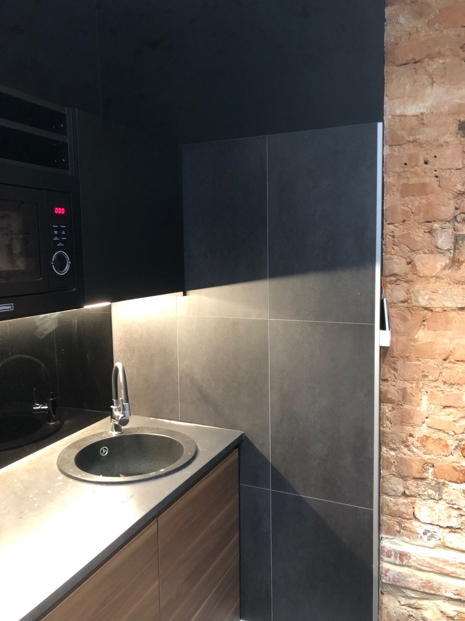 Czarny aneks kuchenny z okrągłym zlewem, baterią, mikrofalówką i fragmentem ceglanej ściany