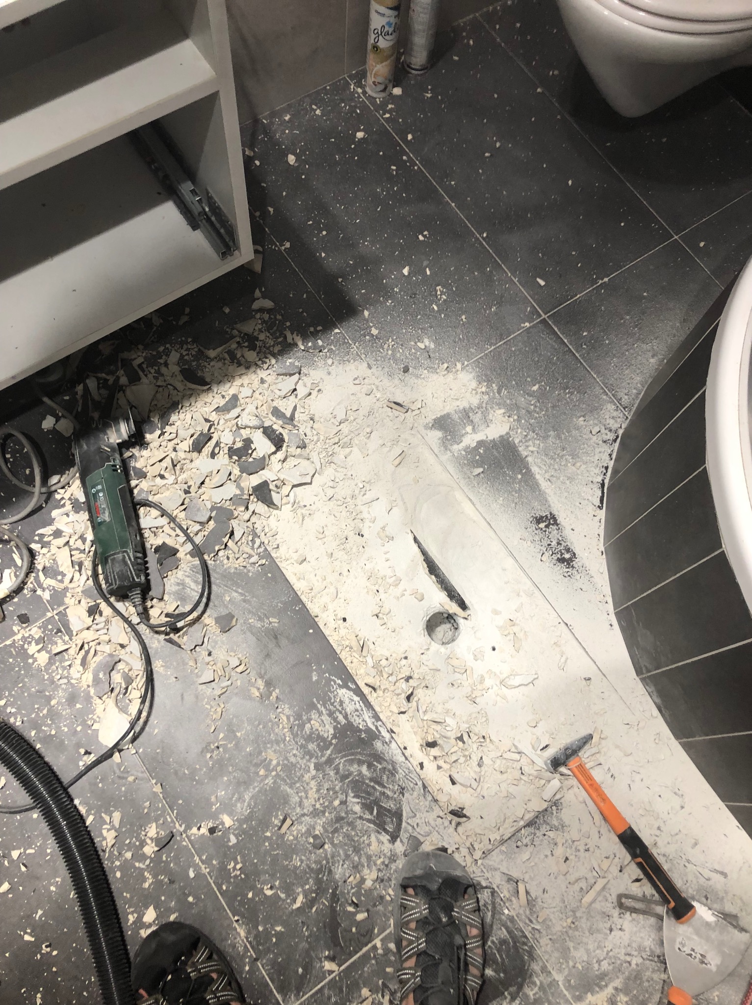 Demontaż obudowy odpływu liniowego w łazience, widoczne narzędzia: młotek, szpachelka i szlifierka, gruz po skuciu płytek, fragment szafki pod umywalkę i toaleta w tle, widok z góry na podłogę...