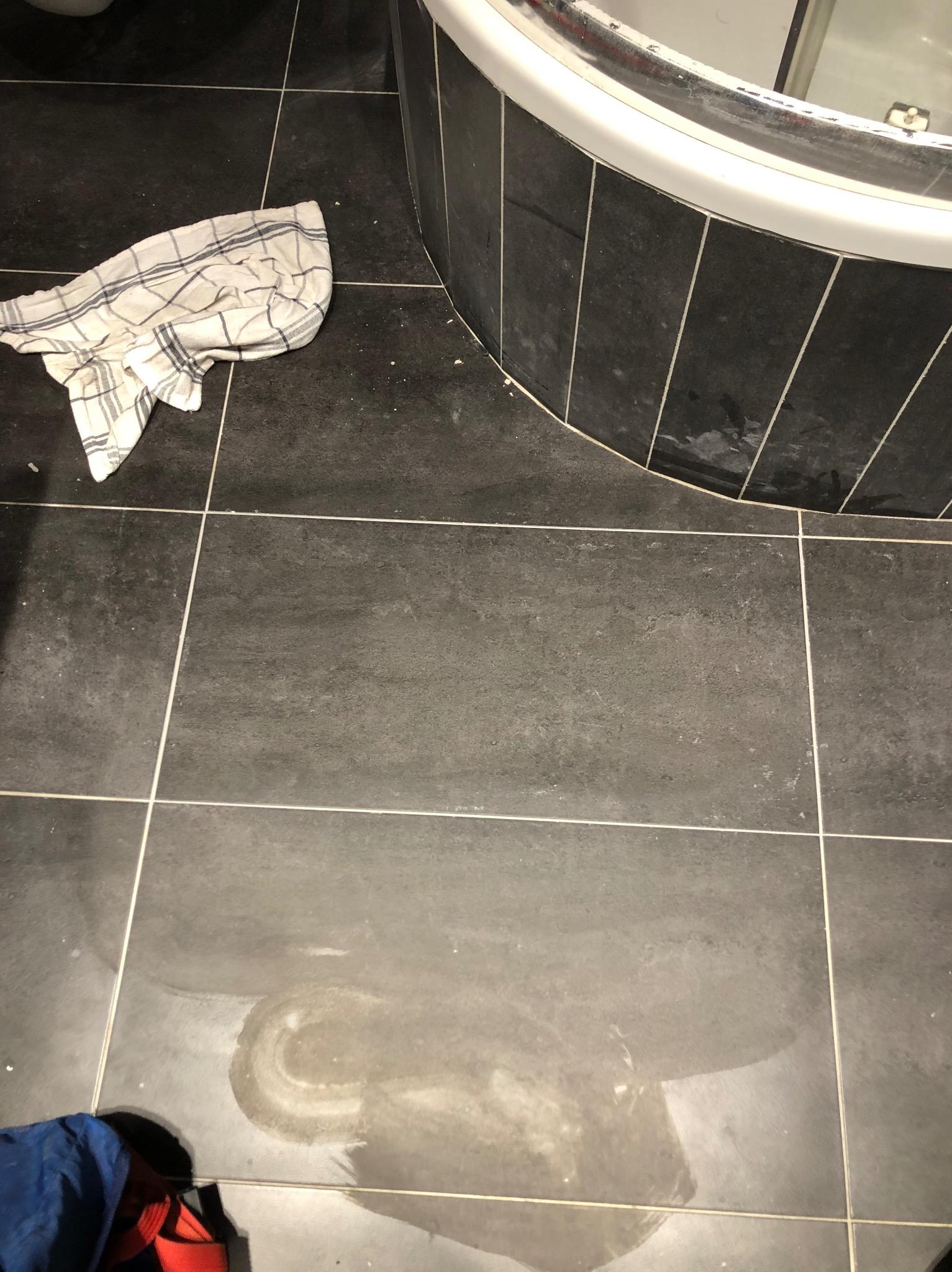 Czyszczenie ciemnych płytek podłogowych w łazience, widoczny ręcznik w kratę, ślady po wodzie oraz fragment wanny.