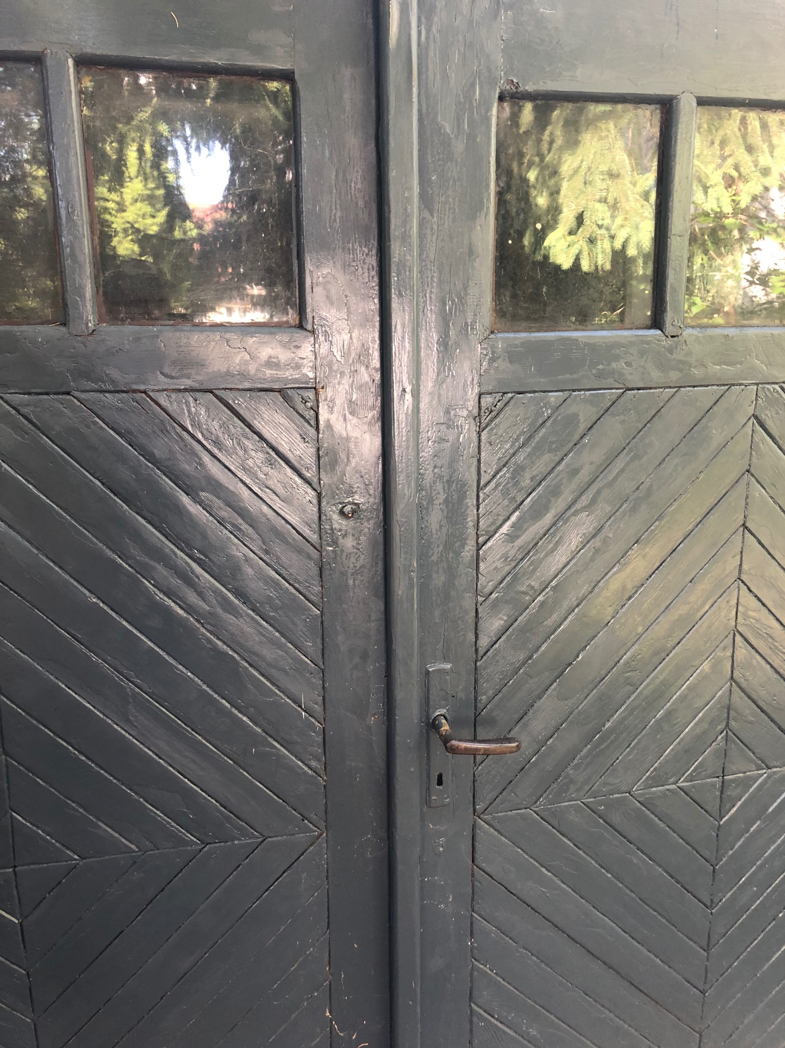 Szare, drewniane drzwi z ukośnym wzorem i małymi okienkami, przez które widać zieleń drzew